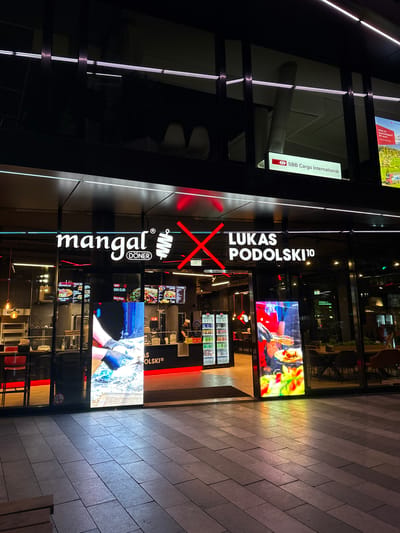 The image shows the exterior of a restaurant called "Mangal Döner Lukas Podolski 10" located in Duisburg, Germany. The restaurant features a prominent illuminated sign with the name of the establishment. To the left of the entrance, there is a large digital display showing a close-up of someone preparing food. Inside the restaurant, a person in a chef's uniform can be seen working behind a counter. The facade of the building is modern, with large glass windows and architectural lighting. The area in front of the restaurant is paved with large square tiles. In the upper part of the image, a sign for "SBB Cargo International" is visible.