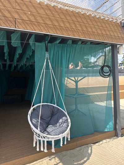 The image depicts an outdoor leisure area in Hlibivka, Ukraine, on a sunny day. A white, circular hanging swing chair with a dark grey padded cushion is centrally positioned in the foreground, suspended by white braided ropes from a black strap attached to a horizontal metal beam. White tassels adorn the bottom edge of the swing chair. The chair rests on a sandy surface, which transitions to a brown wooden deck beneath a shaded structure.

The structure features a ceiling constructed of horizontal wooden planks, partially covered by a white, large-mesh safety net with metal attachments. Teal-colored sheer curtains hang from the structure's framework, defining various sections. To the left, a darker, semi-enclosed section is visible through the curtains, containing a dark object resembling a television screen and a dark, cylindrical pouf.

To the right, another section of the structure is visible through the sheer curtains, revealing a black, four-legged metal table. Beyond the table, two individuals are seated or lounging on a raised surface, partially obscured and blurred by the translucent curtain and bright light. One individual appears to be wearing a red top, possibly swimwear. Their posture suggests relaxation.

The environment outside the structure includes a bright blue sky with scattered white clouds. A white speaker or camera is mounted on a dark grey support beam on the right side of the structure, from which a coiled black cable also hangs. Distant trees and residential buildings are discernible beyond the structure, through the curtains. Sunlight casts distinct shadows, notably from the swing chair onto the sand and from the overhead netting onto the wooden ceiling.