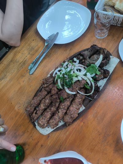 The image shows a wooden table in what is likely a restaurant, given the presence of a meal. On the table are a white plate with a fork and knife, a glass of water, and a basket of bread. The centerpiece is a wooden platter containing a variety of grilled meats, including what appears to be kebabs and other cuts of meat, garnished with sliced onions and herbs. The food rests on what may be thin flatbread. To the lower left, parts of a hand and a green bottle are visible. To the lower right, a dish with a red sauce is partially visible. A person's arm is seen on the top left of the image.

The information about the location being Karatakla, Georgia is not visually verifiable from the image alone. Therefore, I cannot confirm the location.