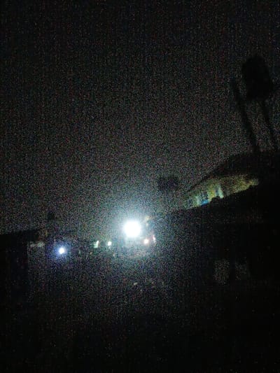 The image, taken at night, features a row of bright lights piercing through the darkness, likely streetlights or building lights. They illuminate a vague structure or building behind them. Above, the dark sky is punctuated with noise, indicating high ISO or low-light conditions. To the right, a dark, undefined shape suggests a tree or pole silhouetted against the night. The overall impression is one of stark contrast between the artificial light and the surrounding gloom.