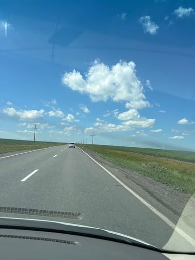 A forward-facing, eye-level shot depicts a car driving on a paved road in a rural landscape under a bright blue sky with scattered white clouds. The road has a white dividing line and is bordered by a grassy shoulder. In the distance, there are telephone poles and a vast green field. A white car is visible driving ahead on the road. The lower portion of the image shows a car's dashboard and windshield. The scene is likely in or near the city of Qazaq, Kazakhstan, given the flat, expansive terrain and typical rural infrastructure often found in the region.