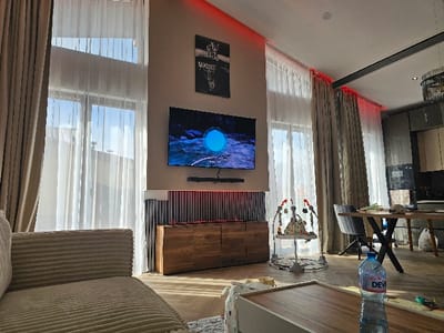 The image displays the interior of a modern, open-plan living and dining area located in Dragushinovo, Bulgaria. The room features light-colored walls and a light wood-patterned floor. Large, floor-to-ceiling windows with both sheer white and opaque beige curtains allow natural light into the space. Red LED strip lighting is integrated along the ceiling edges above the curtain rails.
A wall-mounted television is centrally positioned, displaying a blue circular graphic on a dark background. Below the television is a dark soundbar, and beneath that, a dark wood console cabinet featuring vertical slat details. Above the TV, a framed piece of wall art with white text, possibly "MINUSET," is mounted.
To the left foreground, the ribbed armrest of a light beige sofa is visible. In the midground to the right, a baby bouncer activity center with multiple colorful toys is situated on the floor. Further into the background, a dining area is equipped with a light wood table featuring dark X-shaped legs and dark grey upholstered chairs. Beyond the dining area, a modern kitchen is visible, comprising dark lower cabinetry, integrated appliances including an oven, and lighter upper cabinets.
In the immediate foreground, a light-colored coffee table with a wood-toned top holds a partially full plastic bottle of "DEVI" brand water. No individuals are present within the scene.