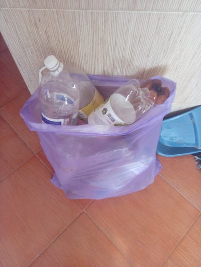 A plastic bag filled with plastic bottles is sitting on a tiled floor next to a wall in an indoor setting. The bag is translucent purple and appears to be slightly crumpled. Several clear plastic bottles with white caps are visible inside the bag. One bottle has a blue and white label. Another bottle has a white label with green and red graphics. A blue plastic dustpan is visible to the right of the bag. The wall behind the bag has a textured, light-colored surface. The floor is tiled with reddish-brown tiles arranged in a diamond pattern. The image does not provide any information to indicate the location in the city of Terebovlia, Ukraine.