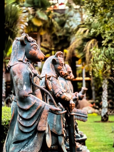 A medium shot captures a series of weathered bronze statues depicting figures with animalistic heads, possibly from the Chinese zodiac, arranged in a garden setting in Vientiane Capital, Laos. The leftmost statue, in the foreground and out of focus, features a cow or ox head with curled horns and a flowing robe. Its right hand is raised in a gesture, and a curved metallic object is held in its left hand. Behind it, other similar statues are visible in decreasing focus, creating a sense of depth. The figures are dressed in traditional attire with ornate details. The background consists of lush green foliage, including palm-like trees and smaller bushes, with hints of a building structure in the upper background, suggesting it is part of a temple or park complex. The lighting suggests it is daytime, with dappled sunlight filtering through the trees. The overall atmosphere is serene and somewhat aged, with the bronze exhibiting signs of patina and wear.