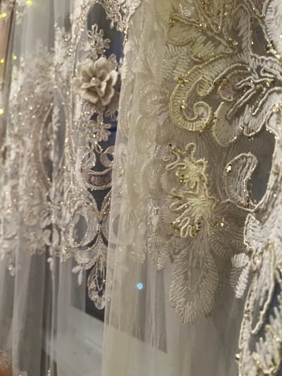 The image shows a close-up of ornate, white lace fabric with intricate floral embroidery and small, sparkling embellishments. The lace is draped, creating layers and revealing a darker background behind the front layer. The fabric appears delicate and possibly decorative, suitable for formal wear or home decor. The scene is static and does not provide any information about a specific location within the city of Мичуринский, Russia.