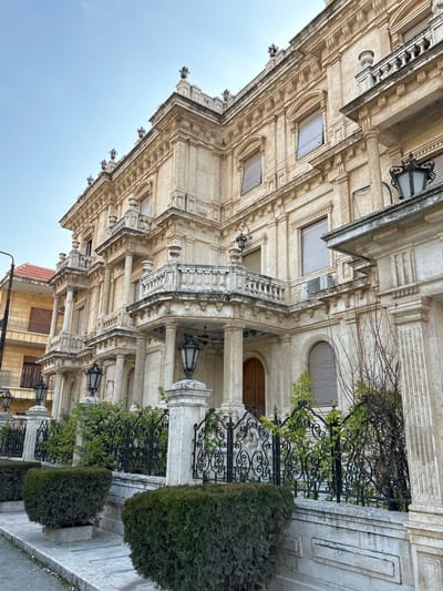 The image displays a grand, multi-story building, likely a mansion, characterized by its ornate classical architecture and light-colored stone facade. The structure is richly detailed, featuring prominent columns, multiple balconies with elaborate balustrades, decorative cornices, intricate window frames, and arched recesses. Several antique-style black metal lanterns are affixed to the building's facade and hang within the covered entrance area, where a large, dark wooden door is visible behind columns.
In front of the building, a meticulously maintained outdoor area is defined by elegant black wrought iron fences with decorative scrollwork, interspersed with neat, dark green hedges. Some sparse tree branches and green foliage are also visible growing near the fences and walls.
The sky overhead is a uniform, pale blue, indicating either an overcast day or diffuse daylight, with no harsh shadows cast. A portion of another, simpler building with a terracotta-tiled roof can be seen in the lower right background. There is no visible activity or text in the image. The setting is in Aleppo, Syria, showcasing architecture that blends traditional regional styles with classical influences.