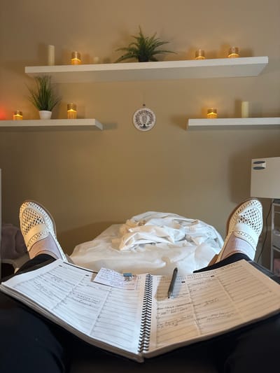 The image shows a first-person perspective looking down at an open planner on a person's lap. The planner contains handwritten appointments and notes. The person is wearing white mesh shoes with pink accents and grey socks. The background consists of a treatment table covered in white linens, two white shelves mounted on a tan wall, and decorative items on the shelves including LED candles and potted plants. A circular wall hanging depicting a tree of life is also visible on the wall. The setting appears to be a spa or wellness center, possibly in Vaughan, Canada, given the context.