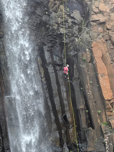 A person is rappelling down a rocky cliff face next to a waterfall. The rappeller is wearing a red and white jacket with the number 8 on the back and a white helmet. A yellow rope is attached to the rappeller and extends upwards, out of the frame. The cliff face is dark and wet in some areas, with patches of brown rock and sparse vegetation. The waterfall is a white curtain of water on the left side of the image. The scene appears to be outdoors in a natural environment, possibly a canyon or gorge. The location is identified as the city of Valok, Russia.