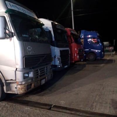 At nighttime in the city of Krasnooktyabrskoye, Russia, a row of semi-trailer trucks is parked. The closest truck, on the left, is silver and features a green sun visor with blue lettering. The second truck is white, followed by a red truck, and then a blue truck on the right. The trucks are positioned on a paved surface, possibly a parking lot or roadside. The background is dark, indicating a lack of significant lighting beyond what emanates from the trucks themselves. There are no people visible in the image. The environment appears to be industrial or commercial, consistent with a truck depot or loading area.