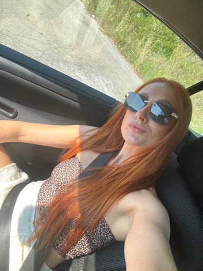 A woman with long, reddish-orange hair and sunglasses is taking a selfie from inside a car, looking towards the camera with a slight smile. She is wearing a leopard-print halter top and white shorts, with a seatbelt across her chest. The background through the car window shows a roadside with greenery and a paved surface. The lighting suggests it is daytime, likely sunny, given the reflections on her sunglasses and the overall brightness of the scene. The interior of the car is visible with black car seats and door trim. The overall mood appears casual and relaxed, possibly indicating a road trip or a leisurely drive.