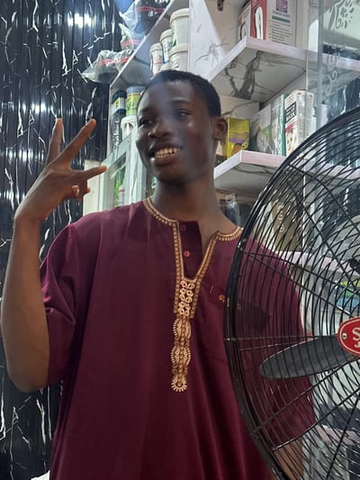 A man stands indoors, partially obscured by a black fan, with retail shelving and items in the background. The man wears a maroon-colored garment with gold embellishments around the neckline. He smiles and raises his left hand in a peace sign. The background consists of shelves displaying various products. Based on the image content, it is not possible to accurately locate the scene in the city of Ogwashi-Uku, Nigeria.