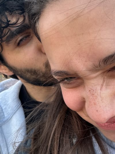 The image shows a close-up of two people, a woman and a man. The woman is positioned in the upper portion of the frame, her face is rotated. She has brown hair, fair skin, and freckles on her nose. The man is positioned in the lower portion, his face obscured and partially out of focus. He has dark curly hair and a beard. Both individuals appear to be close to each other, possibly embracing or lying down. The background is indistinct, but appears to show a white hoodie and black clothing. The location and details relating to Hrazdan, Armenia, cannot be deduced from the provided image.