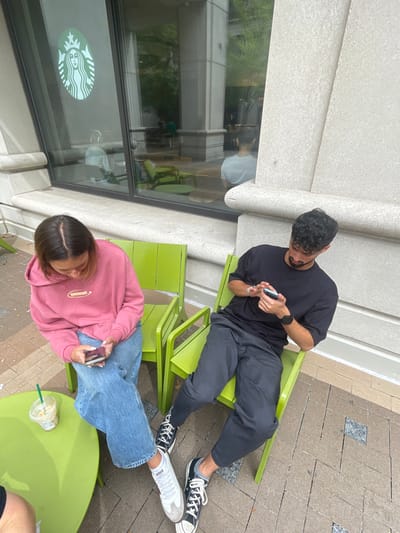 Two individuals are seated in an outdoor patio area outside a Starbucks coffee shop, located in Arlington, United States. On the left, a female wears a pink hoodie with a "BROWNIE" logo, blue jeans, and white sneakers. She is looking down at a white smartphone held in her hands. An iced beverage in a clear plastic Starbucks cup with a green straw rests on a round, lime-green metal table to her right. On the right, a male wears a black t-shirt, dark pants, black and white canvas sneakers, and a dark smartwatch on his left wrist. He is also looking down at a smartphone held in both hands. Both individuals are seated on matching lime-green metal chairs. The background features a large window displaying the Starbucks siren logo, reflecting interior green chairs and the backs of two individuals (one wearing white headphones), as well as exterior trees. The ground is paved with gray and black interlocking pavers.