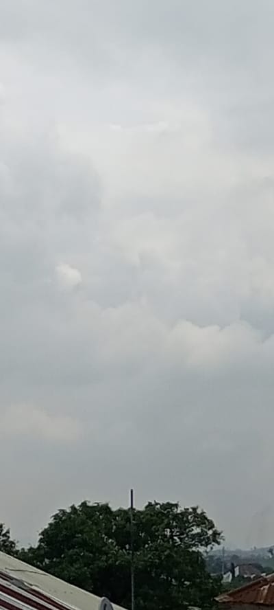 The image depicts an overcast sky with thick, grey clouds filling most of the frame. In the lower portion of the image, a dense, dark green tree is visible, its foliage appearing full and lush. A thin, vertical pole or antenna extends upwards from behind the tree. In the bottom left corner, a portion of a red and white striped roof is visible. To the right of the tree, a hint of a rusted orange rooftop can be seen. The background behind the tree shows a hazy, indistinct landscape that suggests distant buildings and terrain, consistent with an urban or semi-urban environment such as Juwa, Nigeria. The overall scene conveys a sense of cloudy weather.