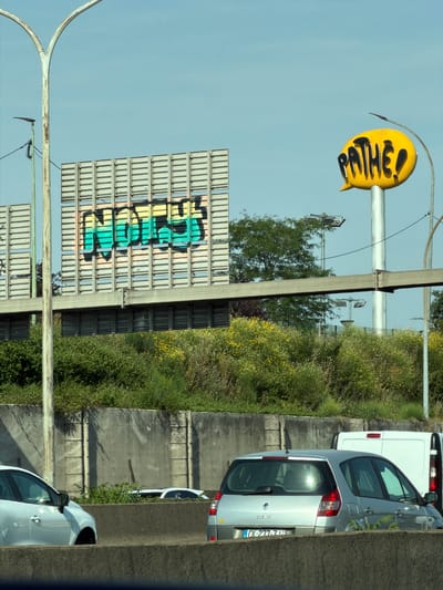 The image depicts a bright, daytime outdoor scene featuring a highway with several vehicles and overhead signage. In the background, a large, grey, slatted billboard displays graffiti in vibrant colors, with the word "NORD" visible. To the right, a yellow speech bubble sign with the word "PATHE!" in black is mounted on a pole. The surrounding environment consists of lush green foliage, including trees and smaller plants with yellow flowers, interspersed with concrete barriers and a light blue sky. Several cars are visible on the road, including a silver Renault Scenic with a license plate reading "DK 277-ZT". A white van is also partially visible. The scene appears to be located on a highway near the city of Rungis, France, indicated by the "PATHE!" signage which is a common cinema chain in France. The overall impression is one of urban infrastructure integrated with natural elements.