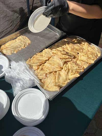 A close-up shot shows a serving of crepes on a table. A person wearing black gloves is holding a white paper plate over a tray of crepes. Another tray of crepes is visible to the left, and a stack of white paper plates and a bag of plastic forks are in the foreground. The table is covered with a dark green cloth, and a shadow suggests the presence of an outdoor setting, possibly near a building or tent. The overall scene appears to be a food service setup.