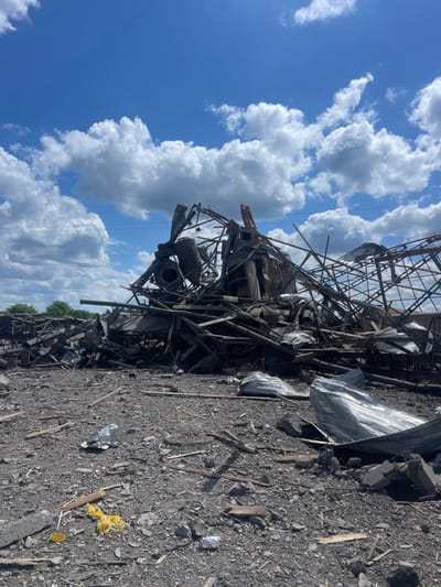 The image shows the remnants of a destroyed building in Terny, Ukraine (latitude: 50.979, longitude: 33.946). The structure, likely an industrial building, is reduced to a pile of rubble and twisted metal. There are no people visible in the image. The debris is scattered across a gravel surface. The sky is clear and blue with white clouds. There is a piece of yellow material on the ground. The scene appears to be the aftermath of a destructive event, possibly an explosion or bombing.