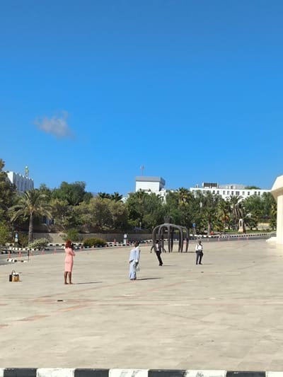 This image captures an outdoor paved plaza in Kızılbaş, Northern Cyprus / Cyprus, under a clear, bright blue sky with a single small white cumulus cloud in the upper left quadrant. The immediate foreground shows a black and white striped curb.
Four individuals are positioned on the light-colored concrete plaza. On the far left, a person with reddish-brown hair, wearing a light pink, short-sleeved dress, stands with their back largely to the viewer, facing towards the right. A black handbag or small luggage case rests on the paved ground to their immediate left. Slightly to the right and further back, a person in a light blue, long-sleeved, floor-length garment holds a light-colored clutch bag in their left hand, also facing right. To their right, a person in dark professional attire is captured in motion, appearing to run or walk briskly from left to right. Further right, a fourth person, wearing a white shirt, dark trousers, and dark shoes, stands stationary, facing right.
In the mid-ground, a prominent dark-colored, abstract sculpture with multiple arching supports, resembling an intricate crown or spider-like structure, stands on the paved surface. The edge of the plaza, beyond the individuals, is delineated by a line of red and white striped bollards. Behind these bollards, a dense band of green vegetation, including several tall palm trees and various bushes, separates the plaza from several multi-story white buildings in the background. One building on the far left features a distinctive green emblem on its facade. Another building in the center background displays a flat gray roof with a communication antenna.