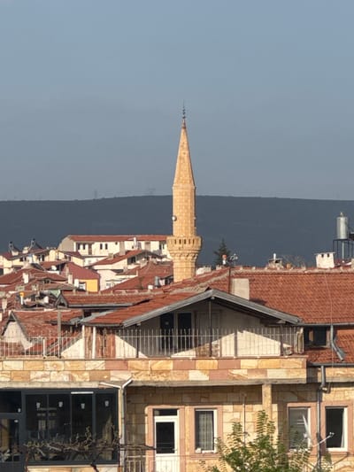 The image depicts a street scene in Ürgüp, Turkey. A tall, slender minaret, constructed of light tan stone, is prominently featured against a backdrop of a hazy blue sky and a dark-colored mountain or distant hill. Adjacent to the minaret is a dense arrangement of buildings, primarily residential, with red tile roofs. The architectural style is compact and layered, typical of urban settings. A building with exposed brickwork and windows with white frames is seen towards the right. Metal scaffolding and railings are visible along its exterior. Other buildings feature stone facades and a mix of windows. The overall scene suggests a historic urban area with a blend of old and new architectural elements.