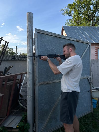 A man is holding a black rifle or long gun in front of a grey metal panel or gate. He is wearing a white short-sleeve shirt, dark shorts, and a watch on his left wrist. He appears to be aiming the rifle, with his head tilted down towards the sights. The environment includes a light blue sky with some clouds, a tree with green leaves, and a building in the background. Below the metal gate, there is a pile of metal items and some vegetation. The location is outdoors in Слобода, Russia.
