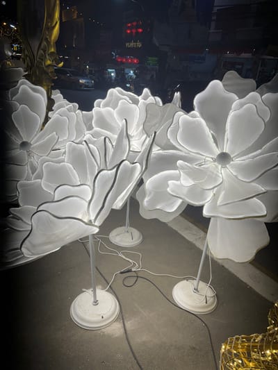 A nighttime scene depicts multiple large, white, illuminated flower sculptures arranged on a dark paved surface in Chiang Rai City Municipality, Thailand. Each sculpture consists of a multi-petaled flower shape crafted from translucent white fabric, internally lit by LEDs, creating a bright glow. These flowers are mounted on white poles anchored by circular white bases. White electrical cords, along with a black one, are visible on the ground, connecting the bases of the sculptures to a power source. In the lower right foreground, a section of a gold-colored, mesh-like illuminated structure is partially visible. The background reveals a street scene with buildings lining the road. A dark-colored sedan is visible on the street, moving away from the viewer. Several signs with Thai script are visible on the buildings, including "ก๋วยจั๊บ" (Guay Jap) and others, some featuring red, circular lantern-like elements. The overall environment is dark, illuminated primarily by the bright flower sculptures and the distant streetlights and building lights. No individuals are present in the immediate foreground.