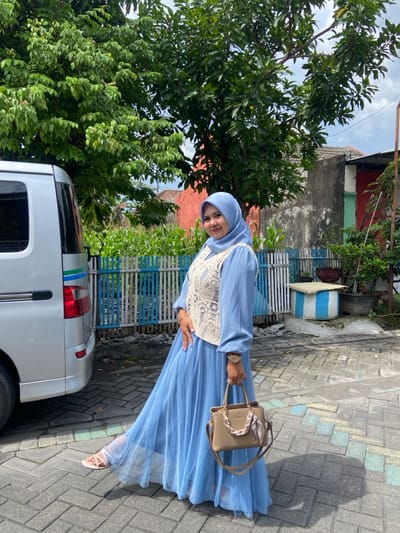 A female subject stands posing on a paved surface in Ketegan, Indonesia. She is oriented slightly right of center, looking towards the camera with a slight smile. She wears a light blue hijab, a light blue long-sleeved dress with a pleated skirt, and a cream-colored crocheted vest. Light-colored sandals are visible on her feet, with her left foot extended forward. She holds a beige handbag, adorned with a patterned scarf, in her right hand. A watch is present on her left wrist and a bracelet on her right. To the left, the rear of a silver van, featuring a vertical blue and green stripe, is partially visible. The foreground ground consists of grey interlocking pavers. In the background, a low wooden fence with alternating light blue and white vertical slats defines a boundary. Beyond the fence, a field of green plants and dense green trees are visible. Various residential structures, including one with a red-orange exterior wall and another with a grey exterior, are present. A blue and white striped cylindrical object is located behind the fence to the right. The scene is observed under natural daylight with a partly cloudy sky.