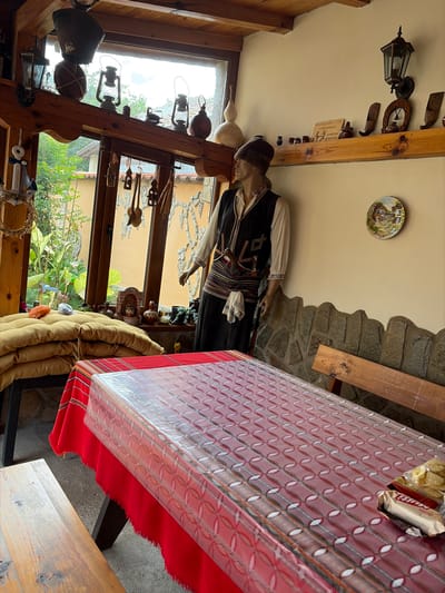 The image shows the interior of a building, likely a restaurant or tourist attraction in Kozlovets, Bulgaria. There's a mannequin dressed in traditional Bulgarian clothing standing near a window. The window offers a view of a garden. A table covered with a red and white patterned tablecloth takes up a significant portion of the frame. On the table are snacks. The room is decorated with various artifacts, including lamps, decorative plates, and wooden carvings, giving it a rustic, cultural feel. The walls are a mix of plaster and exposed stone.
