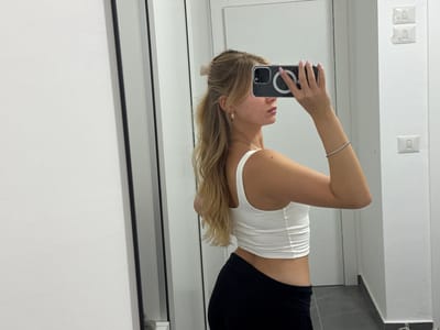 A young woman with blonde hair pulled back in a high ponytail, with bangs framing her face, is seen from the side and rear, taking a mirror selfie. She is wearing a white, ruched crop top and black leggings. She has a gold hoop earring in her left ear and a thin diamond bracelet on her right wrist. Her nails are painted pink. She is holding a smartphone in her right hand, which is reflected in the mirror. The background consists of a white wall with two light switches, and a tiled floor. The overall environment appears to be an indoor space, possibly a bathroom or changing room, with clean white walls and neutral flooring. No specific location in the city of Ariel, Palestinian Territories, can be identified from the visual information provided.