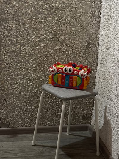 A red, crab-shaped toy keyboard with blue eyes and yellow appendages sits on a gray stool. The stool has thin white legs and is placed on a dark gray wooden floor. The background consists of a textured wall with small, light-colored pebbles and a white, rough-textured wall on the right. The lighting suggests an indoor environment, possibly a room in Астроміцкі сельскі Савет, Belarus.