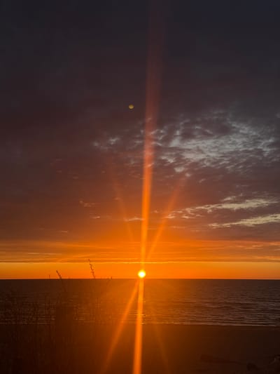 The image shows a sunset at Lilaste beach in Latvia. The sun is setting over the sea, casting a bright orange light across the water and sky. To the left of the sun, there is a dark, clouded sky. To the right is the beach with vegetation. A strong lens flare is visible in the image. The photo is taken vertically.
