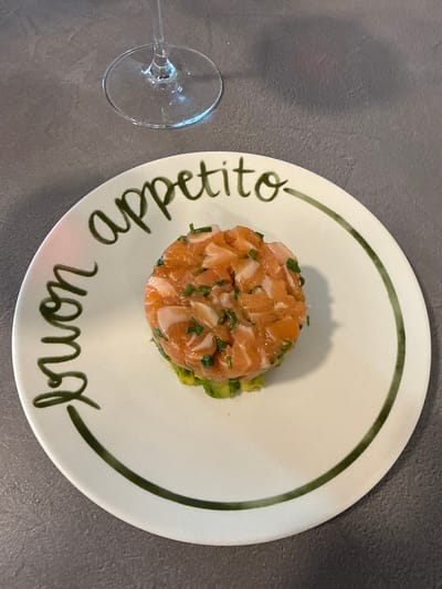A close-up, top-down view shows a salmon tartare served on a white plate with a green rim. The tartare is a mound of finely diced salmon mixed with herbs, placed atop a layer of mashed avocado. The plate is decorated with dark green handwritten text in a cursive script, which reads "Appetito Buon" on the lower half of the plate. The plate is positioned on a dark gray, textured surface, possibly a countertop. To the left of the plate, a portion of a clear wine glass is visible, with its stem and base resting on the same surface. The lighting suggests an indoor setting, possibly a restaurant or home kitchen. There are no people visible in the image. The overall impression is a beautifully presented appetizer, ready to be enjoyed. The text on the plate is Italian, meaning "Good appetite."