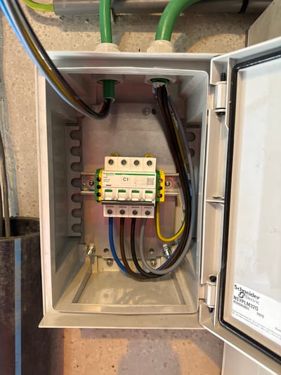 The image shows the interior of an open electrical distribution box. Inside, a row of four Schneider Electric circuit breakers is mounted on a DIN rail. Various colored electrical wires, including yellow/green (ground), blue (neutral), and brown (live), are connected to the circuit breakers and pass through cable glands into the box. The box itself is made of white plastic, and its open lid reveals a black rubber seal. A label on the inside of the lid reads "Schneider Electric NSYPLM32G 4508843891 2620". The electrical box is mounted on a textured, light-colored wall, with a section of dark material with blue stripes visible to the left of the box. The scene is illuminated, suggesting it is an indoor or sheltered outdoor location. No people or specific actions are depicted, the image is a static shot of the electrical equipment. The location is identified as Kallo, Belgium.
