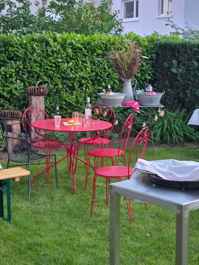 The image depicts an outdoor seating area in Bettembourg, Luxembourg. A bright pink metal table and four matching chairs are arranged on a lush green lawn. On the table, there are several glasses, a small candle in a glass holder, and a plastic cup. A bottle of wine and a blue bottle are also visible. Behind the table, a metal serving table holds two large galvanized tubs, a pitcher with flowers, and several bottles. Decorative light bulbs hang from strings above this serving table. The scene is framed by a dense, green hedge. In the foreground, a portion of a metal and wood bench is visible. A metal food pan covered with a white cloth is placed on a separate silver metal table in the right foreground.