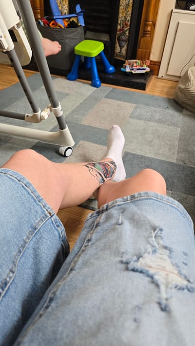 The image shows a close-up of a person's legs, seated on a carpeted floor. The person is wearing ripped denim shorts and white socks. A colorful tattoo of what appears to be a ripped-open section of skin with intricate designs underneath is visible on the left leg, around the ankle area. In the background, to the left, a fireplace with decorative tiles is visible, along with some children's toys, including a bright green and blue plastic stool and a blue toy airplane. A gray laundry basket is also present, containing more toys. The scene appears to be indoors, likely a living room or playroom. The lighting suggests it is daytime. No text is visible in the image.