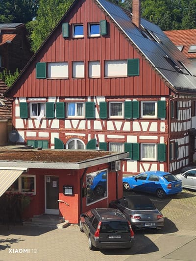 The image captures a sunny day scene of a multi-story building with a red-painted upper section and white-and-timber-framed lower sections, likely a restaurant or guesthouse in Constance, Germany. Solar panels are visible on the steep, red roof. The building has numerous green shutters on its windows. In the foreground, a red-colored single-story structure, possibly an extension or part of the establishment, has an open doorway with stairs leading up to it. Several parked cars are present: a blue hatchback, a dark-colored sedan, and a dark station wagon. The ground is paved with cobblestones. The overall impression is a well-maintained building in a residential or commercial area.