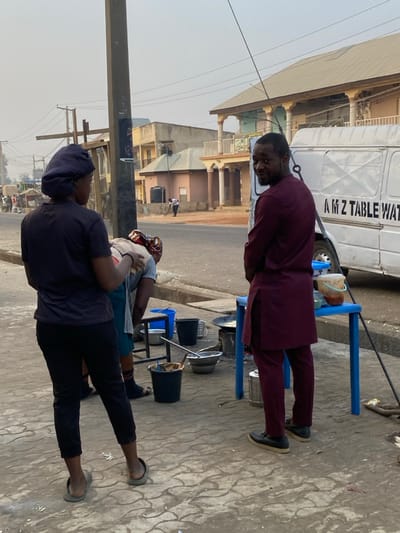 A man in a maroon suit stands outside a van. He is preparing food with several pots, buckets, and metal utensils set up on tables and chairs. Another person in black pants and a dark blue shirt wearing a head covering stands to the side. Both are outside on a paved street with buildings and power lines visible in the background.