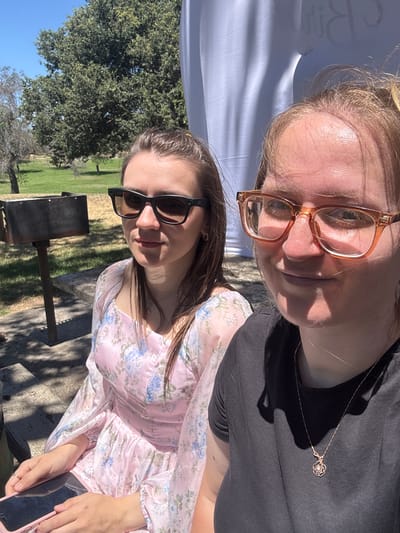 Two women are photographed outdoors. The woman on the left has brown hair and is wearing sunglasses and a pink floral dress. The woman on the right has blonde hair and is wearing glasses and a black t-shirt. She is wearing a rose-shaped pendant necklace. They appear to be sitting at a picnic table next to a grill. The background consists of trees and a white banner. The location is Elverta, California, United States.