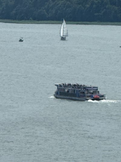 A wide shot of a body of water, likely Lake Mikołajki in Mikołajki, Poland, under an overcast sky. The far bank is lined with a dense dark green forest. In the foreground, a large blue ferry with multiple decks is moving right to left, creating a white wake. The ferry appears to be carrying numerous passengers. Further out on the water, a white sailboat with its sail up is visible in the middle distance, moving right to left. A small, dark boat is also visible in the upper left quadrant of the image. The water is a muted blue-gray, reflecting the cloudy sky.