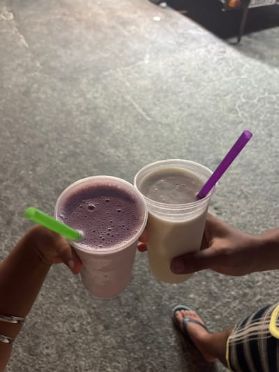 Two people hold up cups of drinks with straws in them. One cup is a light yellow color, and the other is a light purple color. The straws are purple and green, respectively. The drinks are held in the hands of two different people. One is wearing black and white plaid shorts and blue flip flops. There is a concrete floor in the background.
