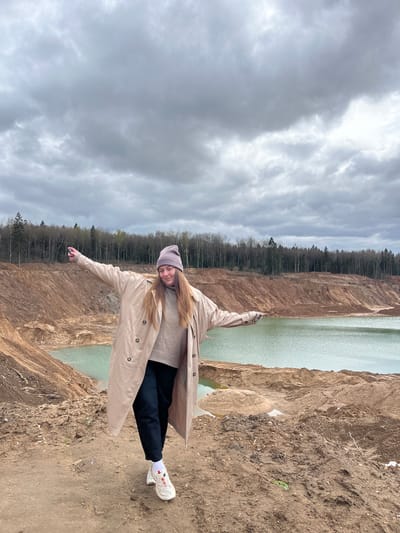 A young woman stands on a dirt embankment, arms outstretched, wearing a tan trench coat, gray beanie, and white sneakers. She is positioned near a quarry filled with greenish water. The quarry is surrounded by dirt cliffs and sparse trees. Above, the sky is overcast with gray clouds. The location is the city of Ivlevo, Russia.