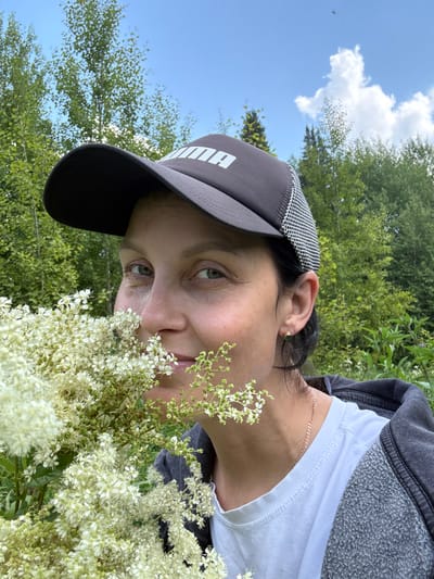 A woman wearing a dark baseball cap with a white logo and a grey zip-up hoodie over a white t-shirt is holding a large bouquet of small white flowers up to her face, partially obscuring it. She has dark hair, dark eyebrows, and green eyes. She is wearing a small gold earring in her visible ear and a delicate gold necklace. The background is a lush green forest with tall trees under a bright blue sky with scattered white clouds. The setting appears to be a natural, outdoor environment, possibly a field or forest edge, in Липовая I, Russia.