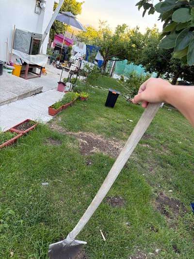 In Golemo Buchino, Bulgaria, a hand holds the wooden handle of a hoe in a grassy outdoor area. The hoe head, a dark metal blade, rests on the ground. Surrounding the hoe is a lawn with patches of disturbed soil. In the background, a white building with external fixtures and a corrugated pipe is visible, along with various garden items including planters, a grill, laundry on a clothesline, an umbrella, and a black toolbox. Trees and vegetation are present, and the sky is clear with daylight.