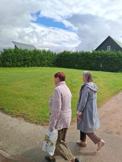 Two women are walking along a gravel path next to a grassy field in Balbieriškis, Lithuania. The woman on the left, with short red hair, is wearing a light-colored jacket and patterned pants and is carrying a white shopping bag. The woman on the right, with gray hair, is wearing a gray raincoat and dark pants. Behind them is a dense hedge and two buildings with dark green, sloping roofs, one of which has a multi-paned window. The sky above is partly cloudy with patches of blue.