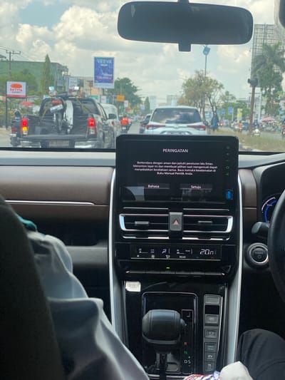 The image captures a view from the interior of a car, looking forward through the windshield and at the car's dashboard display. The car is in motion on a city street in Pemurus Luar, Indonesia. The dashboard display shows a warning message in Indonesian, titled "PERINGATAN," which advises the driver to drive safely, obey traffic regulations, and read the owner's manual for safety information. Below the warning, there are options for "Bahasa" (Language) and "Setuju" (Agree).

Outside the car, the street is busy with traffic. Several cars and a pickup truck carrying a motorcycle are visible. Motorcyclists are also present on the road. Buildings line the street, including commercial establishments with signs. The sky is partly cloudy, suggesting daytime. The interior of the car shows a leather-trimmed dashboard, an air conditioning control panel, and a gear shifter. A person's arm, wearing a light-colored sleeve, is visible on the left side of the frame, resting on the armrest.