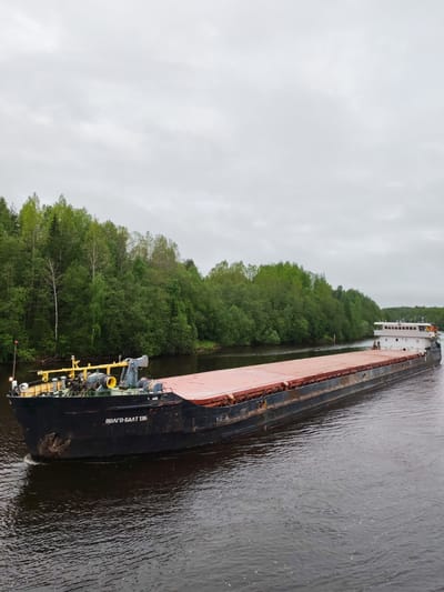 A large barge is moving along a river. The barge is black with a flat, reddish-brown deck. The barge's bow has yellow and gray equipment. The name "ВОЛГО-БАЛТ 136" is written on the side of the barge. The stern of the barge has a white, multi-story structure. The river is dark and reflective. Green trees line the riverbanks. The sky is overcast and gray. The scene suggests a location in Russia, given the Cyrillic text on the barge. The presence of the Volga-Balt waterway system suggests Depo, Russia.