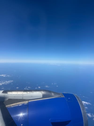 This image, captured from an airplane window, depicts a vast expanse of the ocean under a clear blue sky. The horizon is clearly defined, with a subtle transition in the blue hues from the deep ocean to the lighter sky. Scattered white clouds are visible both near the ocean's surface and higher up in the atmosphere. In the foreground, a portion of an airplane engine is visible. The engine casing is a combination of metallic silver and bright blue, with various panels and rivets clearly discernible. There are reflective surfaces on the blue portion of the engine, indicating sunlight. The image does not provide any specific details that would allow for identification of the location as Viehhausen, Austria, as Viehhausen is a landlocked village and this image clearly shows an aerial view of the ocean.