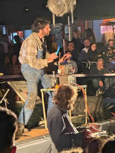 The image shows a man standing on what appears to be a stage, possibly performing or speaking. He is wearing a plaid shirt and jeans with patches. He has a beard and his arms are raised, as if addressing a crowd. In front of him is a keyboard or mixing console. A group of people are in the background, suggesting they are the audience. The lighting seems to indicate an evening or nighttime event. The location, Santa Maria da Feira, Portugal, isn't evident from the image alone. Details that would specify a particular venue or landmark aren't present.