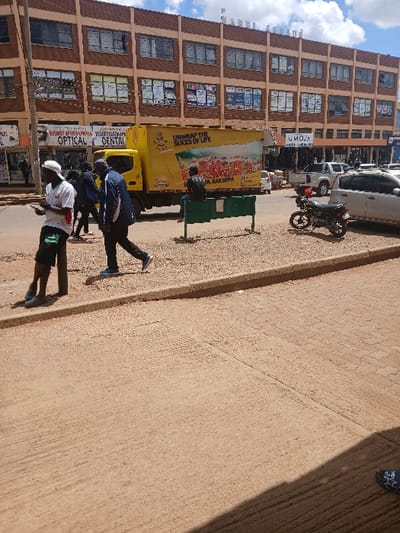 This image depicts an urban street scene in Huruma ward, Eldoret, Kenya, observed during daytime under partly cloudy skies. The foreground features an unpaved, light brown ground surface, likely a sidewalk, separated by a concrete curb from a paved street.

In the left mid-ground, a male individual with a light complexion, wearing a white t-shirt, dark shorts, and dark shoes, stands facing slightly right while engaged with a mobile phone held in his left hand. To his right, another male individual with a darker complexion, dressed in a dark jacket, dark trousers, and blue athletic shoes, walks rightward across the street. Several other indistinct figures are visible walking in the street further back.

A prominent yellow box truck, featuring "UNWRAP THE SLICES OF LIFE" branding, occupies the central mid-ground, oriented to the left. To the right of the truck, a male individual in dark clothing sits on a green metal bench, facing left. A black motorcycle is parked on the right side of the street beyond the bench. Additional vehicles, including a white pickup truck and a white sedan, are visible further down the street.

The background is dominated by a multi-story, light brown commercial building identified as "P. NANDI ARCADE" across its top. Its ground floor hosts various businesses, including "OPTICAL" and "DENTAL" services, and a shop with an "UMOJA" sign. A dark utility pole is visible on the far left.