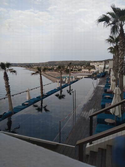 The image depicts a view from a building in Pyli, Greece, overlooking a coastal scene. The foreground features part of a deck or patio with wooden planks and a structure of beams. Blue lounge chairs are arranged along the edge, some with white umbrellas and small tables. Beyond the patio, a narrow strip of sand forms a beach, lined with palm trees. The Ionian sea extends into the distance, and along the coastline, buildings of the town can be seen, nestled at the foot of a hill or mountain range. The sky is overcast with visible cloud formations. Palm tree reflections are visible in the water.