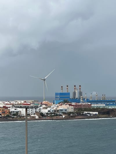 An elevated long shot captures a coastal industrial and residential landscape in Puerto del Rosario, Fuerteventura, Spain, under a heavily overcast, grey sky. In the foreground, choppy, dark blue-green ocean water occupies the lower quarter of the frame. A tall, slender grey pole with a multi-tiered platform topped by a red dome stands vertically in the left foreground, partially obscuring the view of the shore.

The midground features a dense array of residential buildings, predominantly white and pastel-colored, lining a rocky coastline. Several parked vehicles are visible on streets between the buildings. Behind and to the right of the residential area, a large industrial complex, identified as a power plant, dominates the background. It comprises extensive blue and grey structures with multiple tall smokestacks, some emitting faint plumes of white smoke or steam. To the left of the power plant, a prominent white wind turbine with three rotating blades rises above the residential structures. No individuals are discernible within the scene.